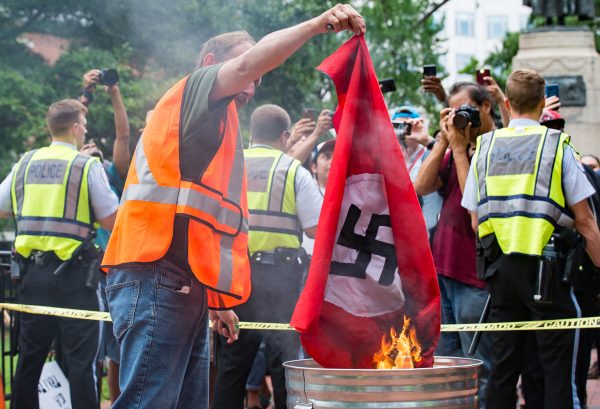 A flag with a confederate battle flag on one side and a swastika on the other is burned by anti-Unite the Right 2 protesters. (Mike Jordan/BPE)